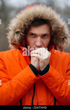 Porträt eines jungen gefrorenen Mannes in Kapuze und orangefarbener Jacke. Nahaufnahme von erwachsenen männlichen wärmenden Händen mit seinem Atem bei kaltem Wetter Stockfoto