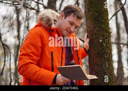 Erwachsene Mann in orange Jacke liest Buch im Herbst Wald. Junger Mann ruht mit Buch an der frischen Luft, genießt seine Freizeit Stockfoto