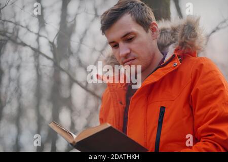 Erwachsene Mann in orange Jacke liest Buch im Herbst Wald. Junger Mann ruht mit Buch an der frischen Luft, genießt seine Freizeit Stockfoto