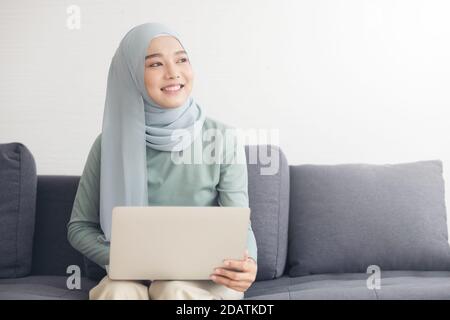Porträt einer jungen glücklichen muslimischen Frau in Hijab arbeiten zu Hause mit Laptop-Computer, während auf dem Sofa sitzen. Stockfoto