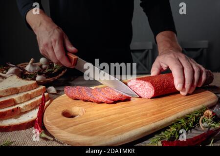 Mann schneidet eine Wurst für ein Sandwich. Geschnittene Salami mit Rosmarin, rotem Pfeffer und Knoblauch auf einem alten Holztisch. Stockfoto