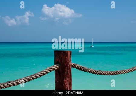 Rustikales Seil mit einem hölzernen Stützstock. Im Hintergrund das karibische Meer mit einem kleinen Segelboot, dem blauen Himmel und weißen Wolken. Mexikanische Karibik Stockfoto