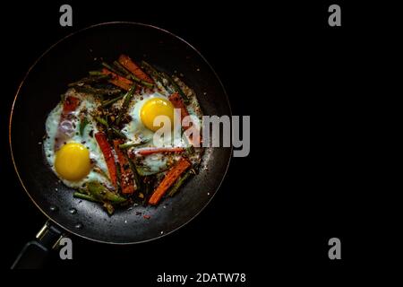 Zwei Spiegeleier in einer Pfanne mit großen Karotten-Scheiben, Paprika und Spargel mit Gewürzen und Sauce. Stockfoto