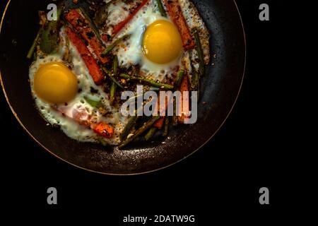 Zwei Spiegeleier in einer Pfanne mit großen Karotten-Scheiben, Paprika und Spargel mit Gewürzen und Sauce. Schließen. Stockfoto