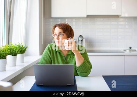 Nachdenkliche ältere Frau mit einer Tasse warmen Tee, die von zu Hause aus mit einem Laptop arbeitet. Sie schaut weg. Stockfoto