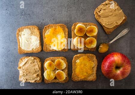 Süßes Frühstück Draufsicht Foto. Toasts mit verschiedenen Belägen: Erdnussbutter, Marmelade, Butter und karamellisierte Banane. Dunkelgrauer strukturierter Hintergrund mit Stockfoto