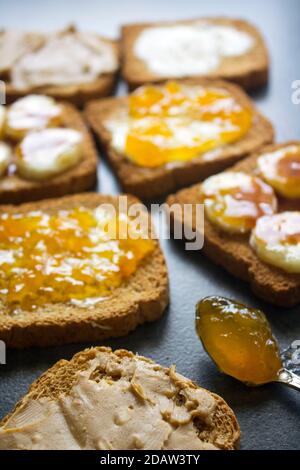 Süßes Frühstück Draufsicht Foto. Toasts mit verschiedenen Belägen: Erdnussbutter, Marmelade, Butter und karamellisierte Banane. Dunkelgrauer strukturierter Hintergrund Stockfoto