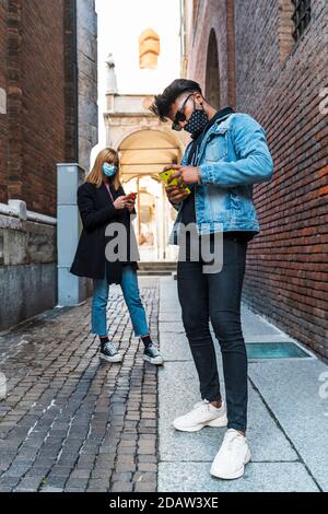 Junge multiethnische Paar mit Schutzmasken mit Smartphones zu teilen Inhalte in sozialen Netzwerken im Freien Stockfoto