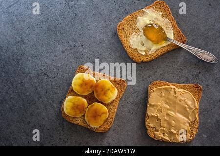 Süßes Frühstück Draufsicht Foto. Toasts mit verschiedenen Belägen: Erdnussbutter, Marmelade, Butter und karamellisierte Banane. Dunkelgrauer strukturierter Hintergrund mit Stockfoto