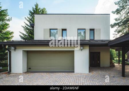 Modernes Äußeres von Luxus-Privathaus mit eingebauter geschlossener Garage. Gefliestes Quadrat. Weiße Wand mit Fenstern. Stockfoto