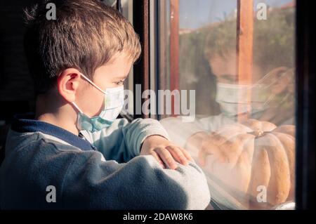 Krankheit Kind in Quarantäne zu Hause. Junge tragen schützende medizinische Masken sitzt auf Fensterbank und schaut aus dem Fenster. Virenschutz, Coronavirus-Pandemie, p Stockfoto