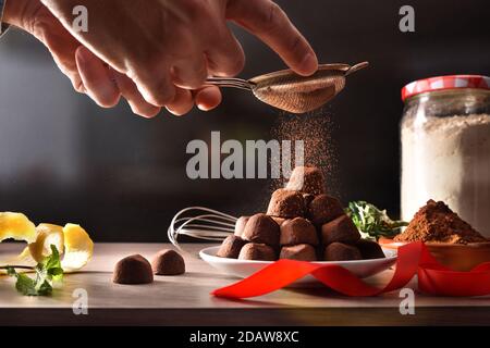 Hände Gießen Kakaopulver auf Stapel von frisch gemachten Schokoladentrüffel auf Tisch mit Band und schwarzem Hintergrund. Vorderansicht. Horizontale Zusammensetzung. Stockfoto