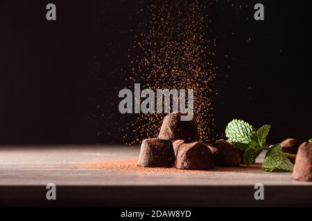 Schokoladentrüffel mit Minzblättern auf Holztisch und dunklem Hintergrund fallendes Kakaopulver. Vorderansicht. Horizontale Zusammensetzung. Stockfoto