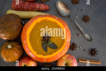 Kürbissuppe in Kürbisschüssel. Draufsicht Foto von Kürbis, Äpfeln, Karotten Anis Sterne und Zimtstangen. Grauer Hintergrund. Ideen für Thanksgiving- Stockfoto