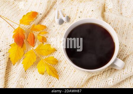 Großer weißer Becher mit heißem, dampfendem Kaffee, Ohrhörern und gelbem Herbstblatt auf einem beigefarbenen Strickschal. Gemütliches Herbststimmung Konzept. Draufsicht. Stockfoto