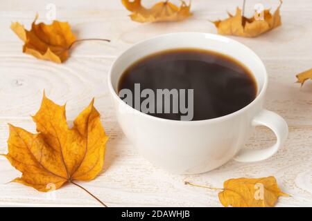 Große weiße Tasse heißen dampfenden Kaffee und einige trockene gelbe Herbstblätter über weißem Holz rustikale Oberfläche. Gemütliches Herbststimmung Konzept. Vorderansicht. Stockfoto