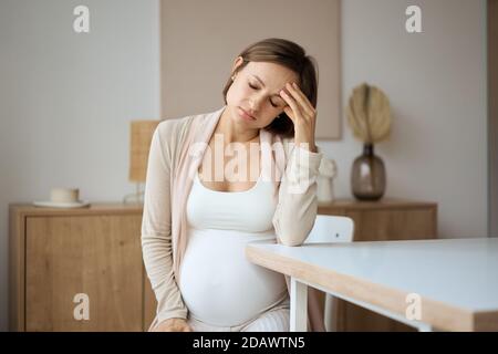 Schwangere Frau, die an Kopfschmerzen leidet, während sie am Tisch sitzt Stockfoto