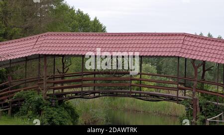 Eine hölzerne Brücke mit zwei hölzernen Karrenrädern und einem Geländer mit einem dunkelroten Metallziegeldach über einem Kleiner Fluss auf dem Hintergrund von grünen Bäumen und Sträuchern Stockfoto