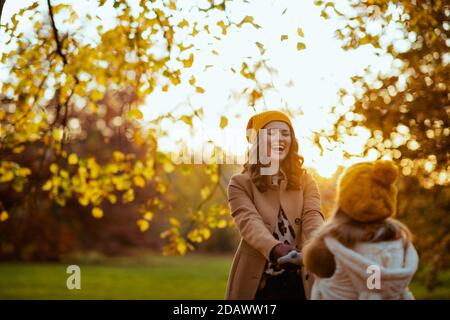 Hallo oktober. Lächelnde moderne Mutter und Kind in gelben Hüten draußen auf dem Stadtpark im Herbst Spaß haben Zeit. Stockfoto