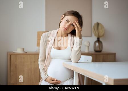 Schwangere Frau, die an Kopfschmerzen leidet, während sie am Tisch sitzt Stockfoto