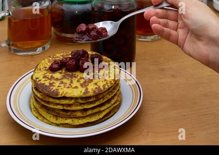 Süßes Frühstück: Frau Hand mit Löffel gießen Kirschmarmelade zu Stapel von frischen heißen Pfannkuchen Stockfoto