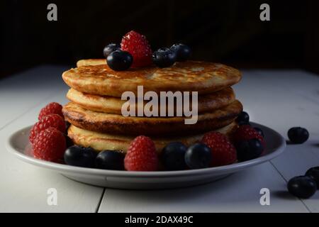 Pancakes mit Ahornsirup, Heidelbeeren und Himbeeren auf weißem Holztisch und schwarzem Hintergrund. Stockfoto