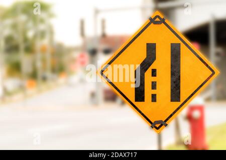 Ein schwarzes Straßenschild auf der gelben linken Lane Ending Straße. Keine Wörter, nur Piktogramm. Geringe Schärfentiefe. Platz für Text. Stockfoto