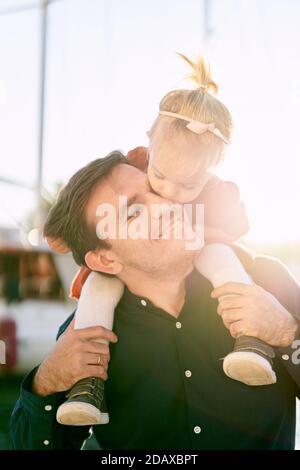 Die 2-Jährige sitzt auf den Schultern ihres Vaters und küsst sich Er auf der Wange Stockfoto