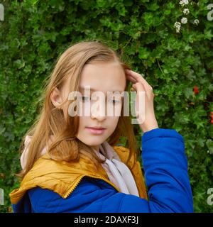 Schöne junge Teenager blonde Schulmädchen in einem blauen und gelben Fell in der Nähe einer blühenden Liane in Form eines Lebende Wand im Park Mitte Herbst Stockfoto