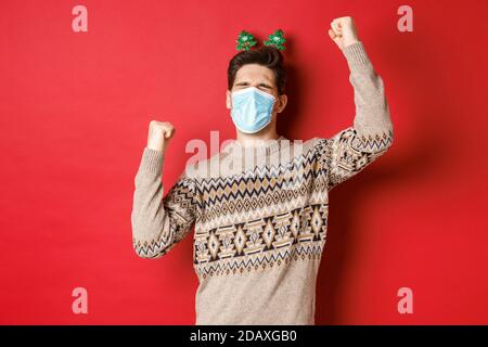Konzept von Coronavirus, Winterurlaub und sozialer Distanzierung. Bild von aufgeregt und glücklich Mann in medizinische Maske und weihnachtskleidung, die Hände nach oben zu heben Stockfoto
