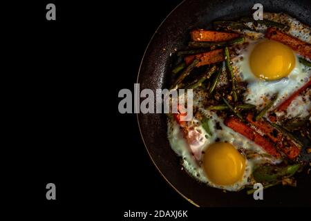 Zwei Spiegeleier in einer Pfanne mit großen Karotten-Scheiben, Paprika und Spargel mit Gewürzen und Sauce. Schließen. Stockfoto