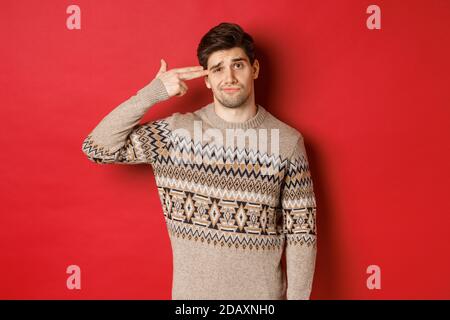 Portrait von verzweifelten und verärgerten schönen Mann, trägt weihnachts-Pullover, Herstellung von Fingerpistole Zeichen in der Nähe des Kopfes und schießen sich selbst, stehend über rot Stockfoto