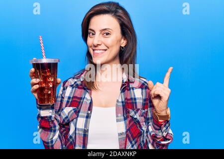 Junge schöne Frau trinkt Glas Cola Getränk lächelnd glücklich Mit Hand und Finger zur Seite zeigen Stockfoto