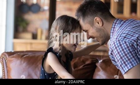 Lachendes kleines Mädchen, das auf dem Sofa sitzt und den Kopf zum Vater lehnt Stockfoto