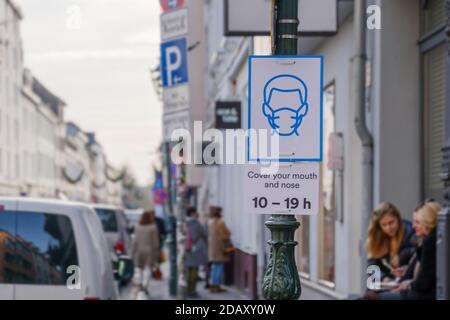 Selektiver Fokus auf schützende Gesichtsmaske Beschilderung auf Pfosten auf Bürgersteig in Deutschland, um Menschen in der Stadt Maske während Epidemie COVID-19 zu suggerieren. Stockfoto