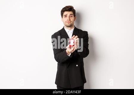 Porträt eines gutaussehenden Mannes, der einen Vorschlag macht, ihn zu heiraten, Ring zeigt, in schwarzem Anzug vor weißem Hintergrund stehend Stockfoto