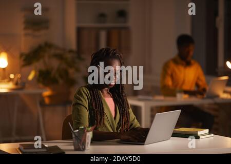 Porträt einer zeitgenössischen afroamerikanischen Frau, die einen Laptop benutzt, während sie spät im dunklen Büro arbeitet, Kopierraum Stockfoto