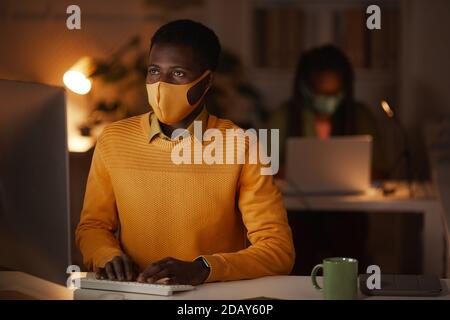 Porträt eines zeitgenössischen afroamerikanischen Mannes, der im Büro eine Maske trägt, während er spät in der Nacht arbeitet, beleuchtet von Laptop-Licht, Kopierraum Stockfoto