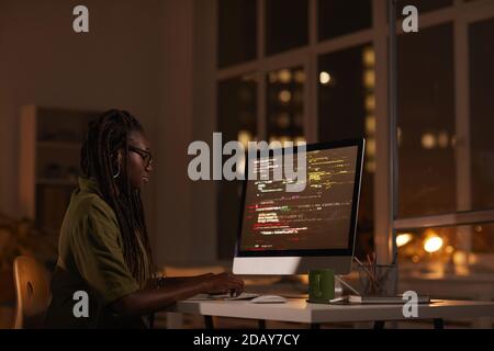 Seitenansicht Porträt einer zeitgenössischen afroamerikanischen Frau, die auf den Computerbildschirm schaut und Code schreibt, während sie spät in der Nacht arbeitet, Platz kopieren Stockfoto