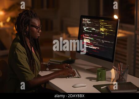 Seitenansicht Porträt einer zeitgenössischen afroamerikanischen Frau, die Code schreibt und auf den Computerbildschirm schaut, während sie im dunklen Büro arbeitet, Kopierraum Stockfoto