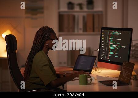 Seitenansicht Porträt einer zeitgenössischen afroamerikanischen Frau, die Code schreibt und auf den Computerbildschirm schaut, während sie in einem dunklen Büro arbeitet, Kopierraum Stockfoto
