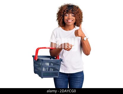 Junge afroamerikanische Frau hält Supermarkt Warenkorb lächelnd glücklich und positiv, Daumen nach oben tun ausgezeichnet und Zustimmung Zeichen Stockfoto