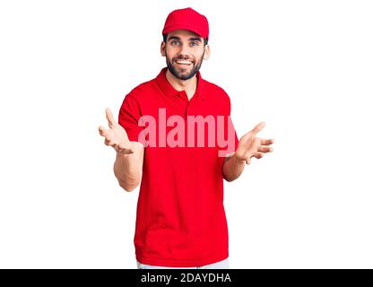 Junge gutaussehende Mann mit Bart trägt Lieferung Uniform lächelnd fröhlich mit offenen Armen als freundlicher Empfang, positive und zuversichtlich Grüße Stockfoto