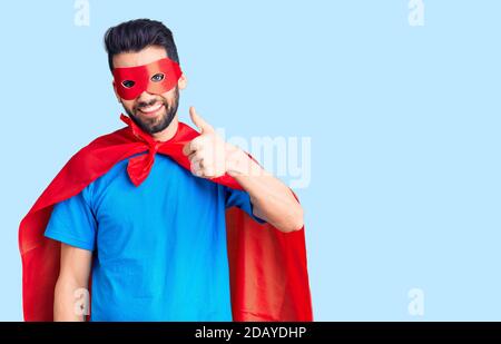 Junger schöner Mann mit Bart trägt Superhelden Kostüm tun glückliche Daumen nach oben Geste mit der Hand. Genehmigung Ausdruck Blick auf die Kamera zeigt s Stockfoto