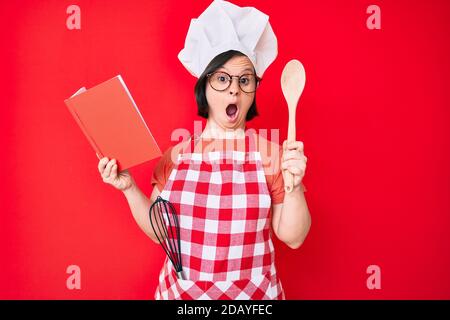 Brünette Frau mit Down-Syndrom tragen professionelle Bäcker Schürze lesen Kochen Rezept Buch Angst und schockiert mit Überraschung und erstaunt Ausdruck, Stockfoto