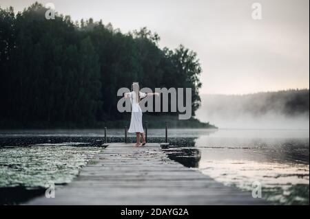 Rückansicht einer jungen Frau in weißem Kleid, die allein auf dem Steg steht und auf den See starrt. Neblig kühl Morgen mit einem Nebel über Wasser. Stockfoto