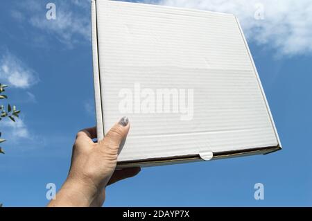 Eine Frauenhand hält eine weiße Pappschachtel mit einer Kopie des Raumes und des Platzes für Text auf einem blauen Himmel Hintergrund, Schablone, mockup Stockfoto