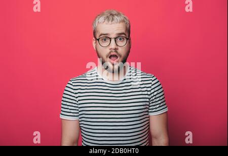 Der blonde Mann ist erstaunt über etwas, während er die Kamera anschaut Durch Brillen an einer roten Studiowand Stockfoto