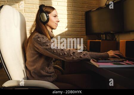 Junge Frau in Kopfhörer mit Videoanruf über Computer zu Hause. Remote-Arbeit, Sonnenlicht an der Wand. Leerer Bildschirm für Ihr Foto. Stockfoto
