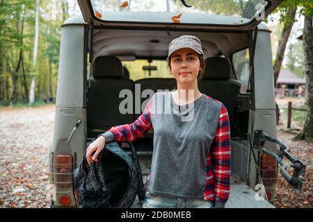Eine junge kaukasische Frau posiert mit einem Rucksack gegen den offenen Kofferraum eines Jeeps. Im Hintergrund Bäume und Wald. Nahaufnahme. Konzept der Autofahrt A Stockfoto